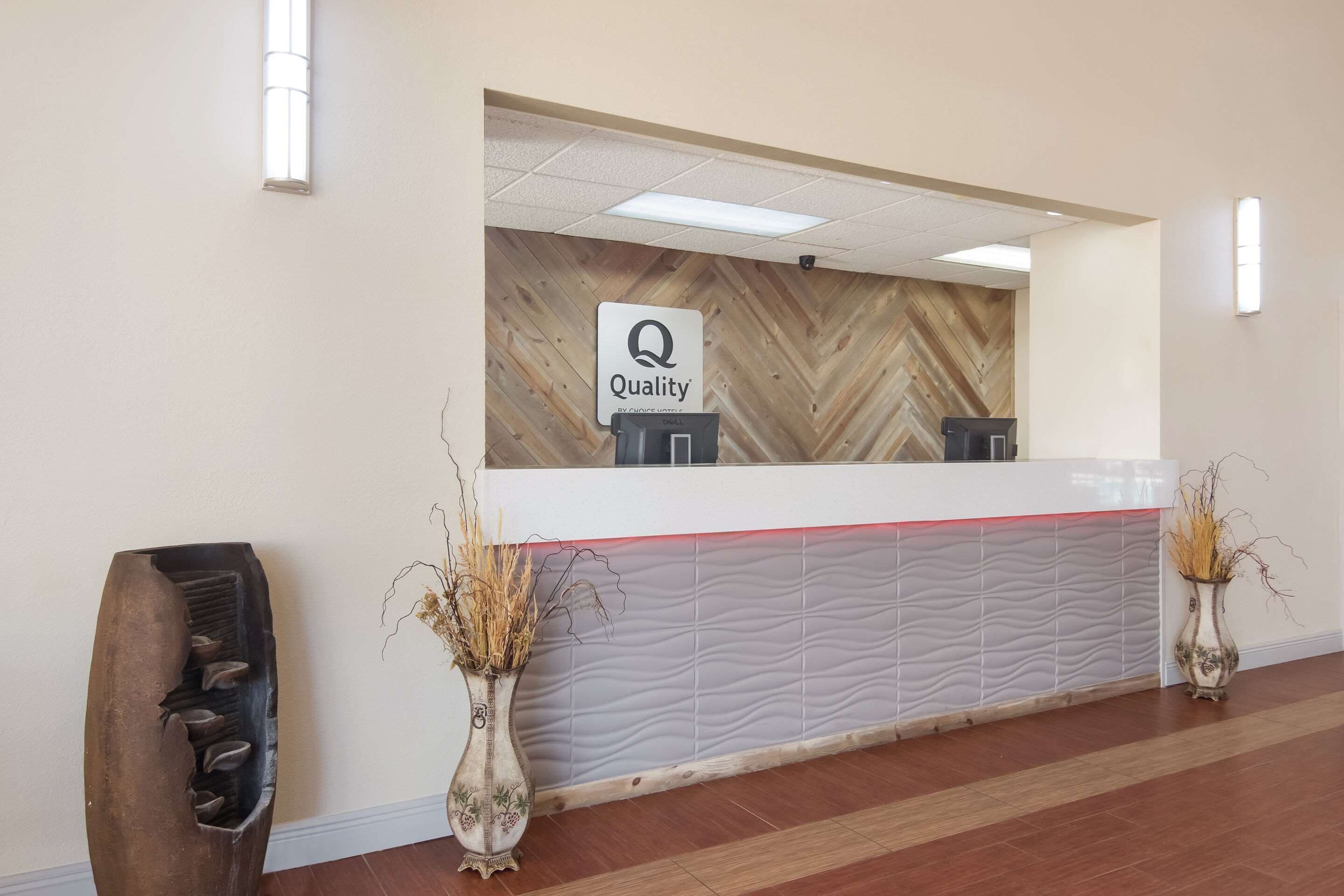Un mostrador de recepción con un cartel que dice 'Quality', una pared con motivo de espiga y dos jarrones con plantas secas.