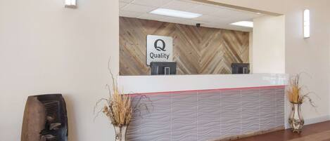 Un mostrador de recepción con un cartel que dice 'Quality', una pared con motivo de espiga y dos jarrones con plantas secas.