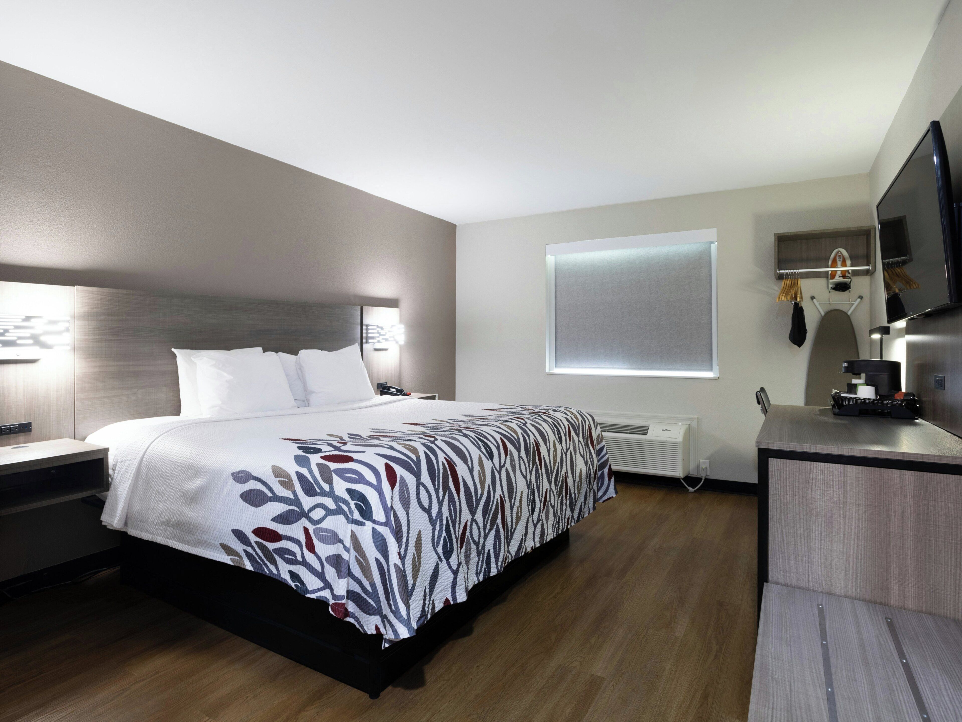 Habitación de hotel moderna con cama, mesita de noche, lámpara y un pequeño escritorio con cafetera.