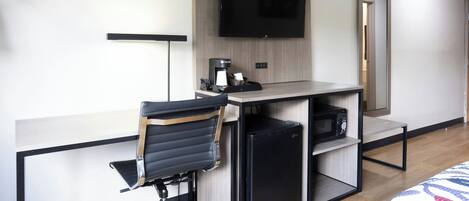 A hotel room with a desk, a chair, a television mounted on the wall, a coffee maker, and a microwave.
