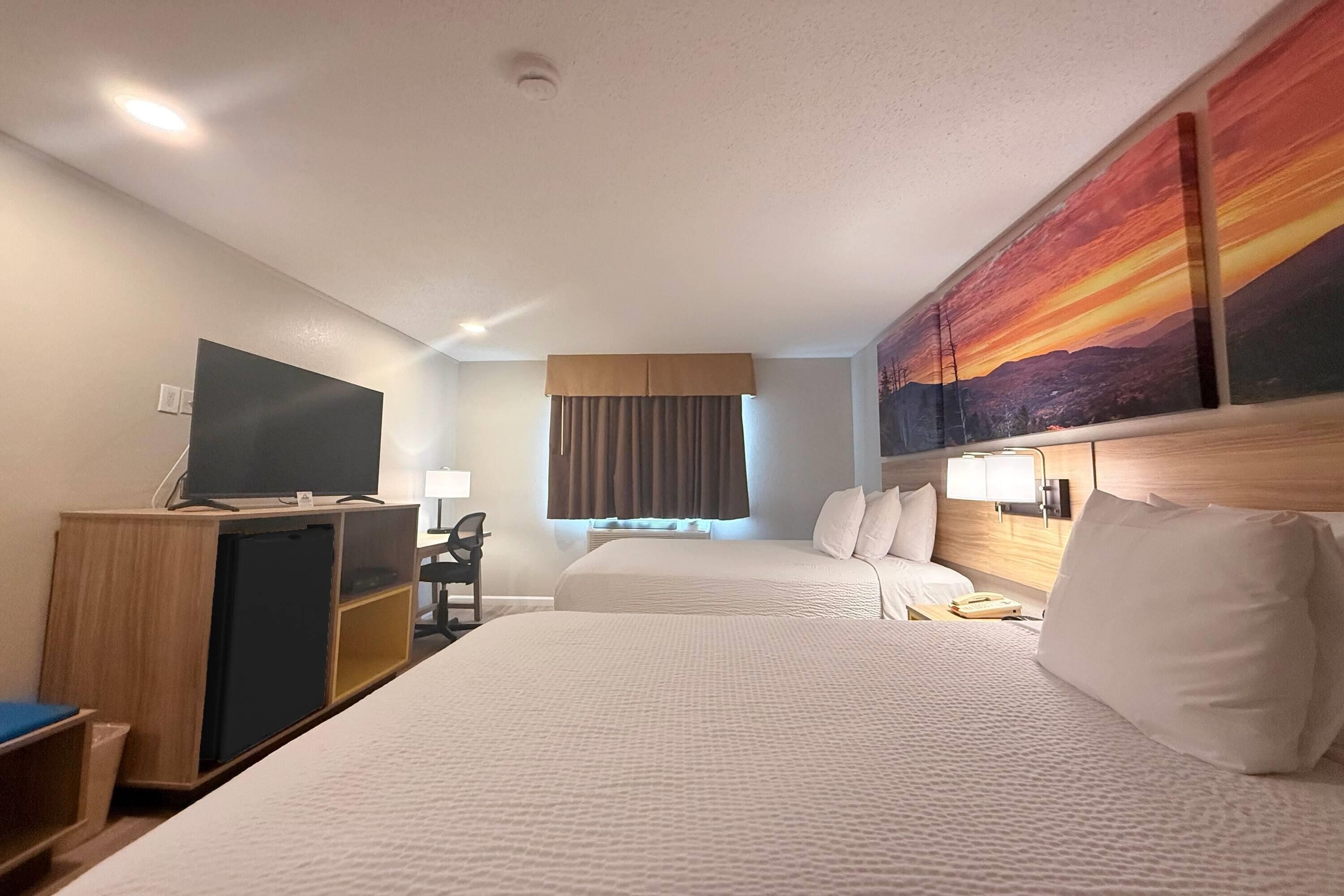 A hotel room with two beds, a TV, a desk, and a painting of a sunset.