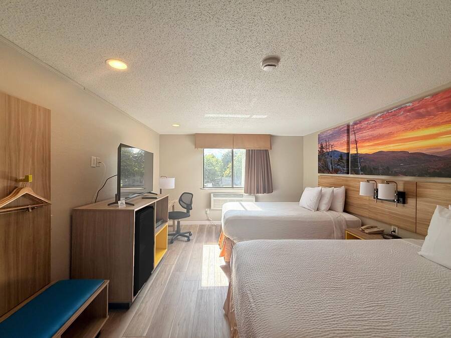 Habitación de hotel con dos camas, un escritorio con computadora, una silla, un banco, un televisor y un cuadro de un atardecer.