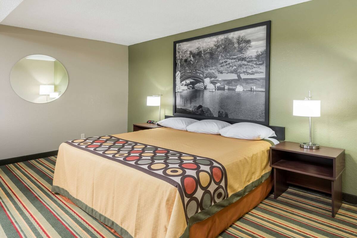 A hotel room with a bed, a round mirror, a nightstand, a lamp, and a framed picture on the wall.