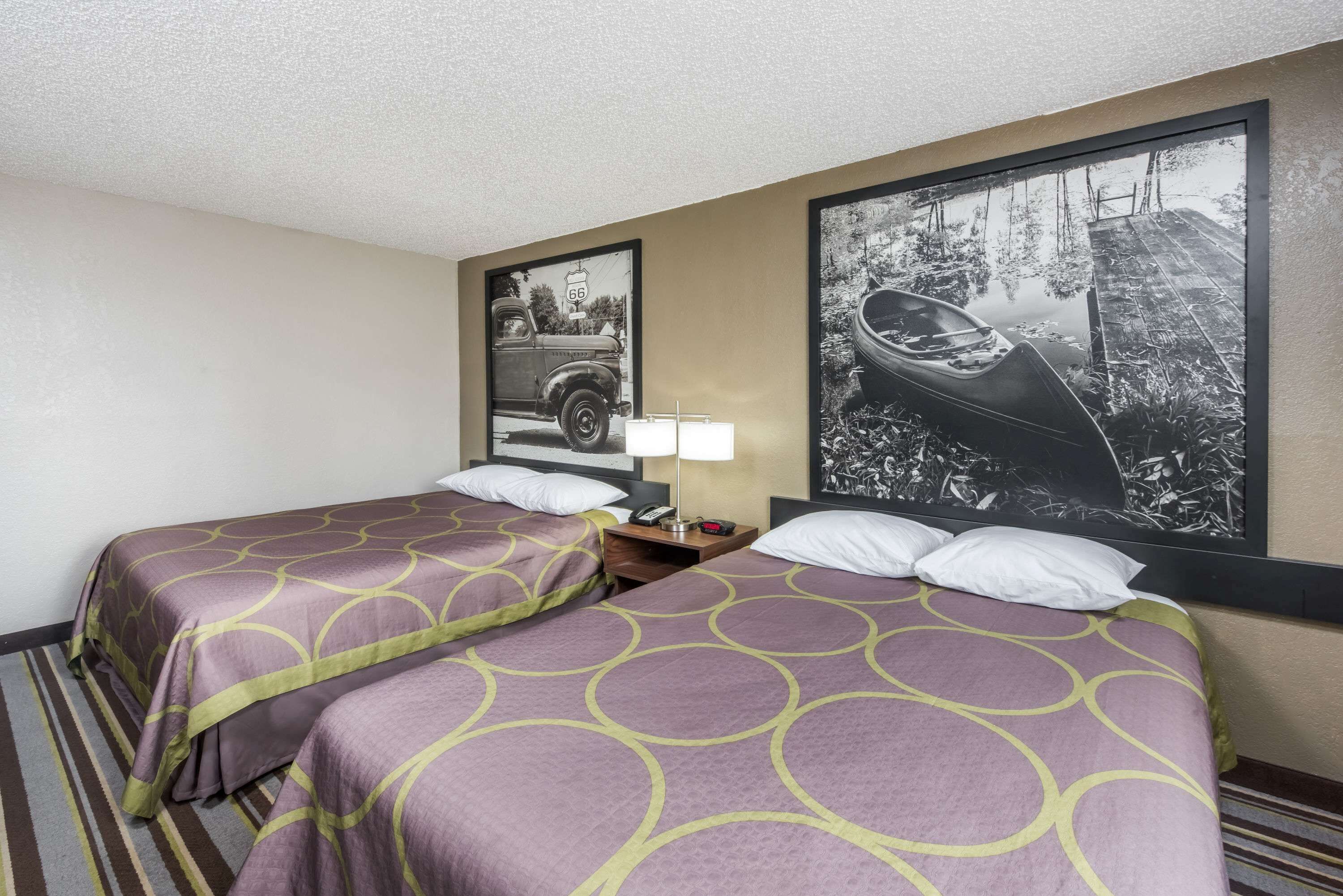 A hotel room with two beds, a nightstand, a lamp, and two framed pictures on the wall.