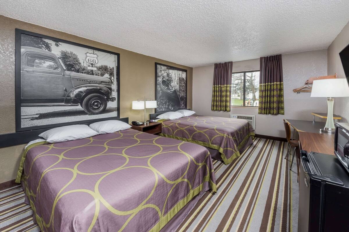 A hotel room with two beds, a large poster of a vintage car, a window with curtains, a desk with a lamp, and a television.