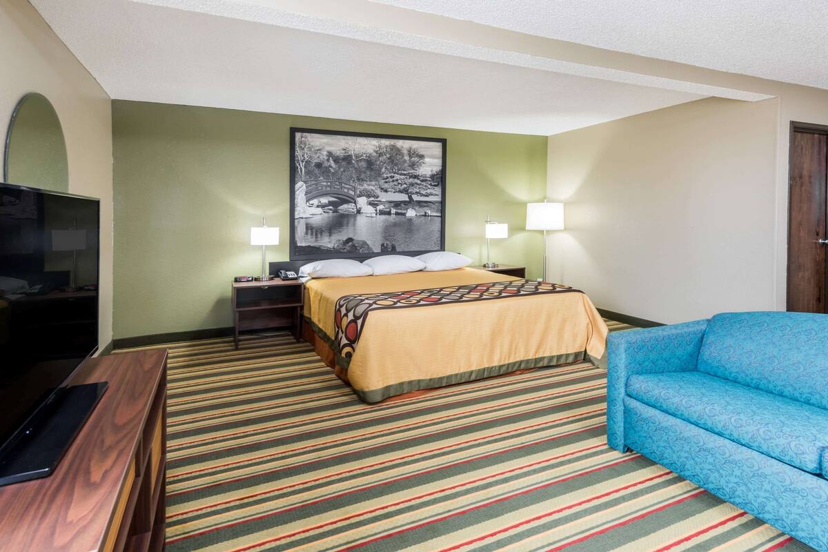 A hotel room with a bed, a blue sofa, a television, and a painting on the wall.