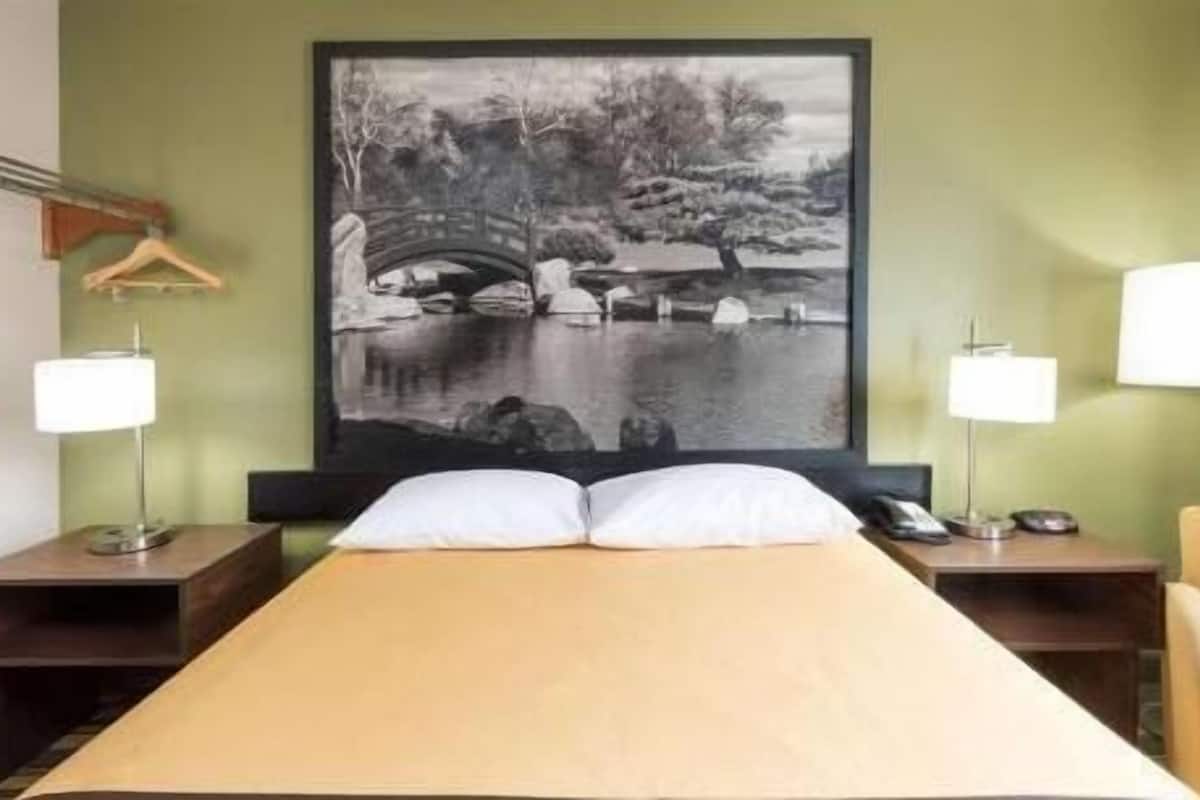 A hotel room with a bed, two bedside tables, a lamp, a chair, and a large black-and-white painting of a bridge over water.