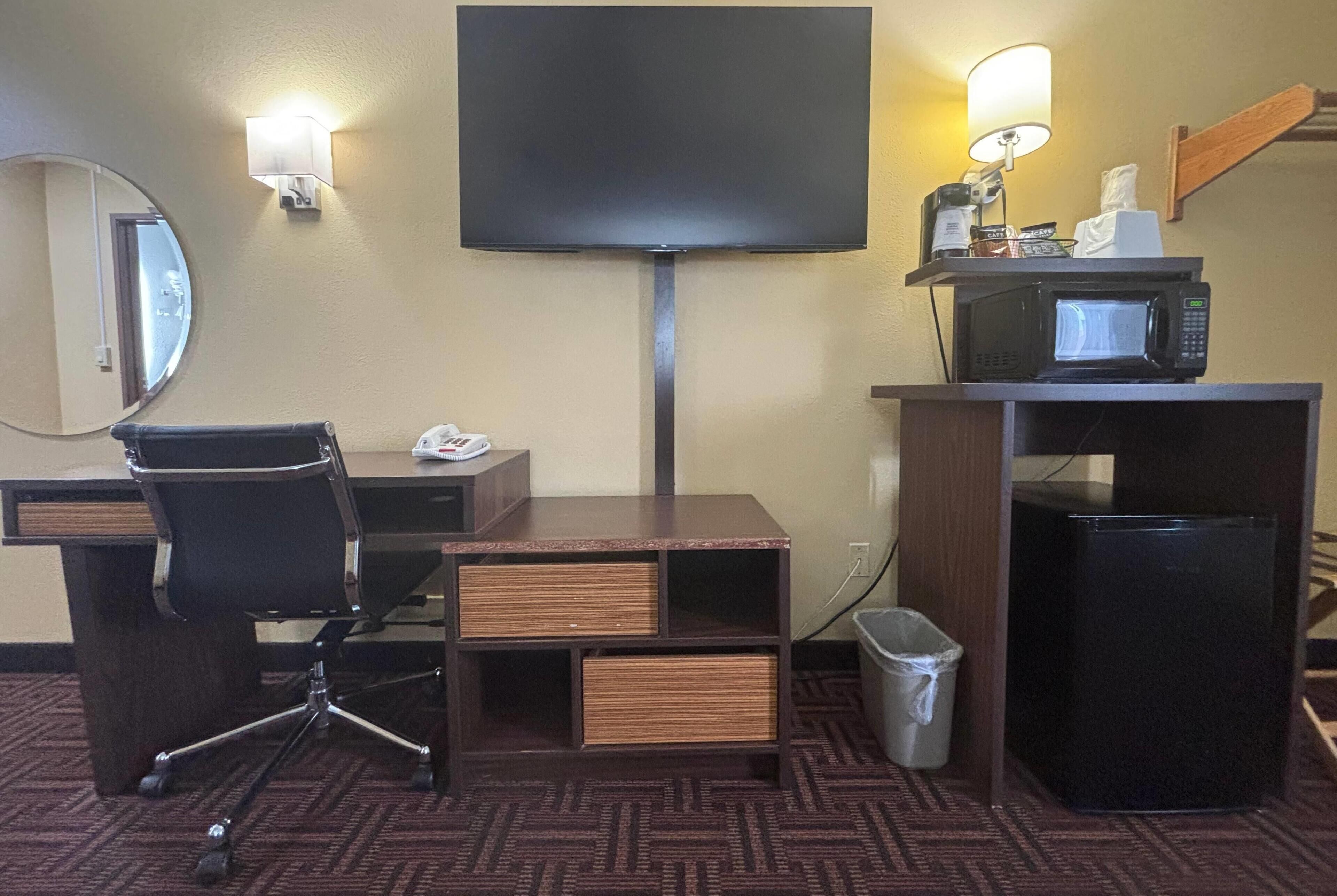 A hotel room with a desk, chair, television, microwave, and trash can.