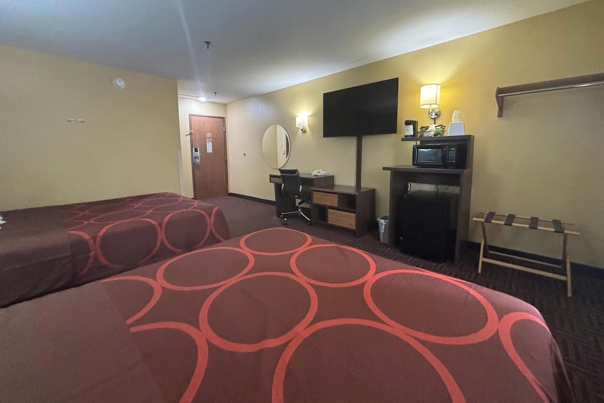 Une chambre d’hôtel avec deux lits, un bureau, une chaise, une télévision, un four à micro-ondes et une petite table avec une lampe.