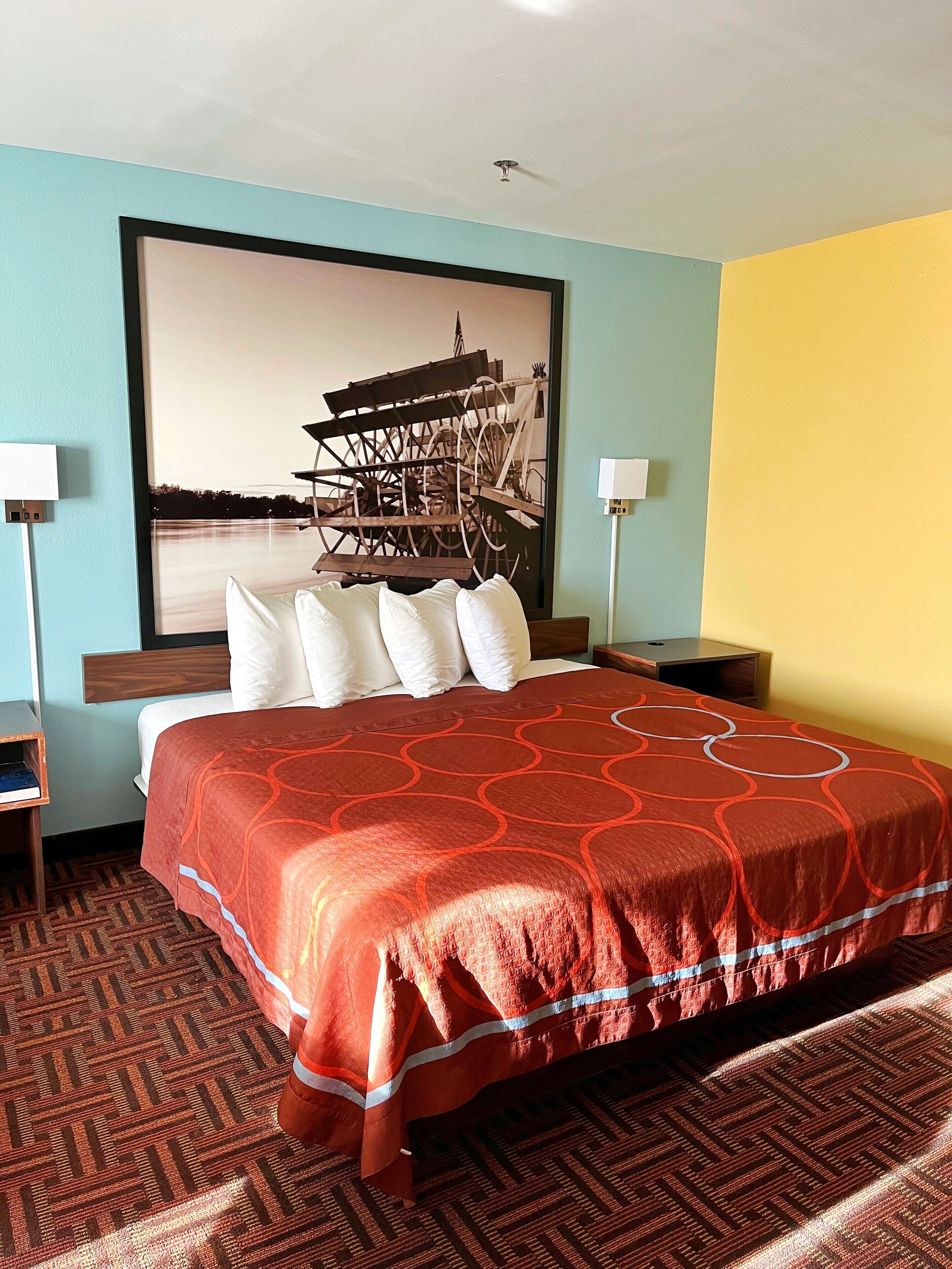 A hotel room with a large bed, a headboard with multiple pillows, a nightstand with a lamp, and a wall-mounted picture of a water wheel.