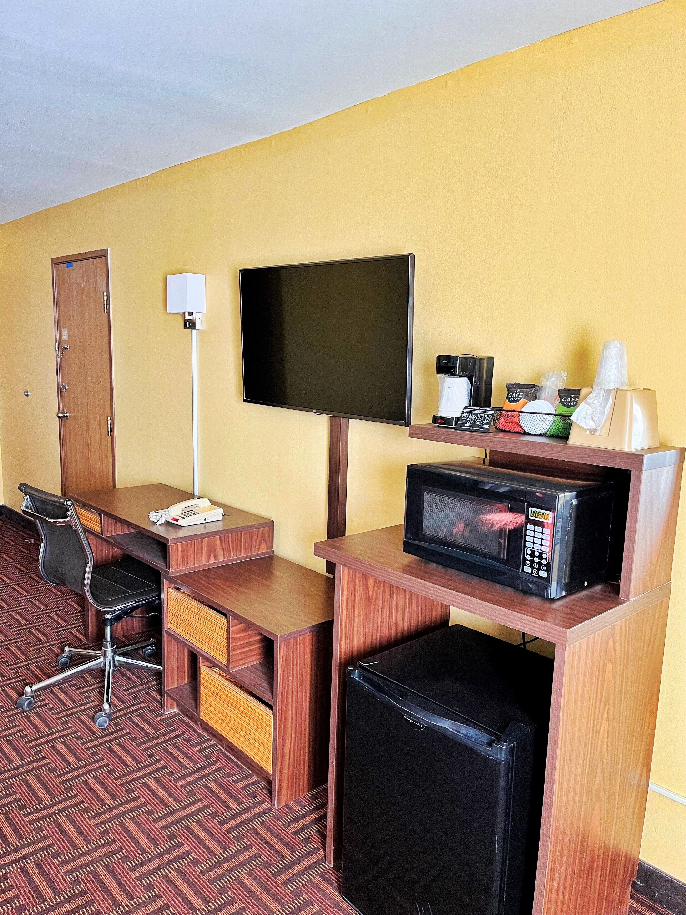 Habitación de hotel con escritorio de madera, una silla, televisor, microondas, mini refrigerador, puerta y repisa con artículos de aseo.