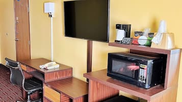 Habitación de hotel con escritorio de madera, una silla, televisor, microondas, mini refrigerador, puerta y repisa con artículos de aseo.