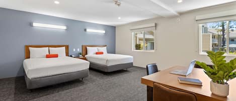 A hotel room with two beds, a desk with a laptop, a potted plant, and a window with a view of buildings.