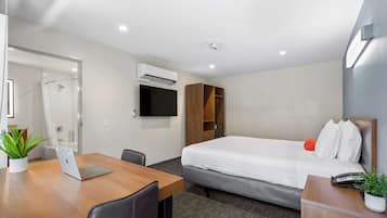 A hotel room with a bed, a desk with a laptop, a chair, a TV mounted on the wall, and a small plant on the desk.