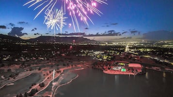 Vista aérea noturna de uma cidade com fogos de artifício, um lago e uma cadeia montanhosa ao longe.