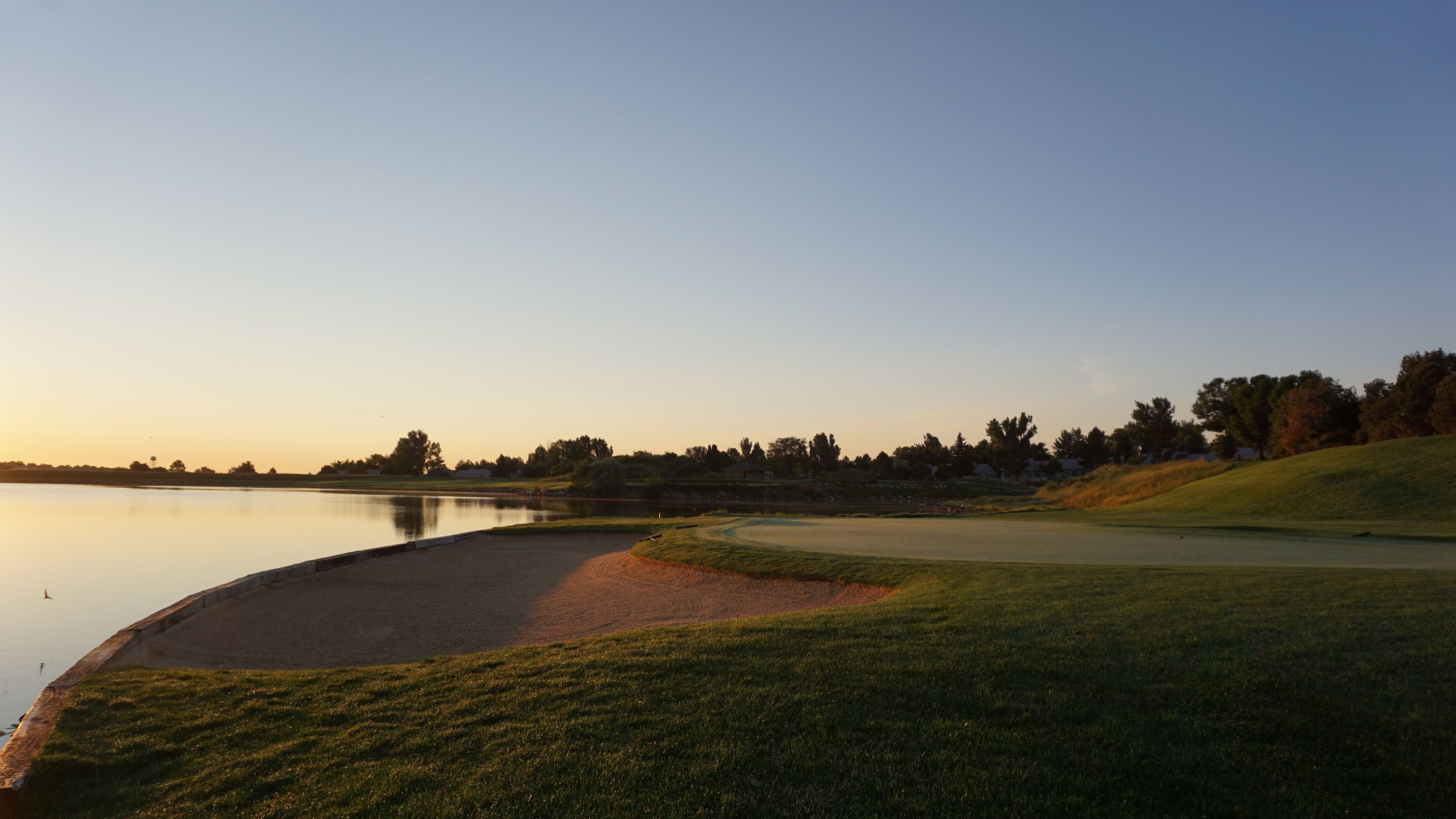 Un campo de golf con un estanque, zonas verdes y árboles al fondo.