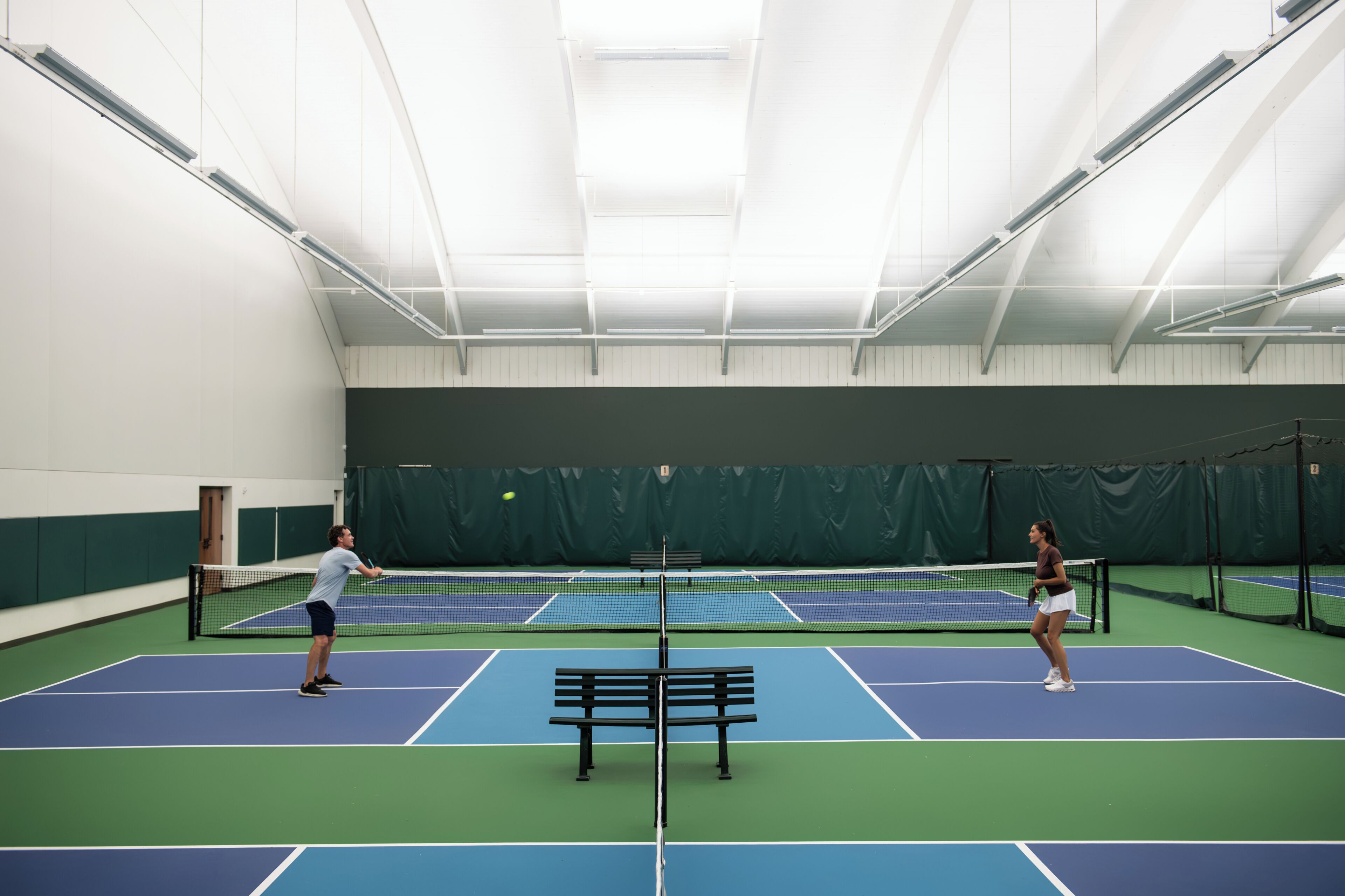 An indoor tennis court with a net, benches, and a green wall.