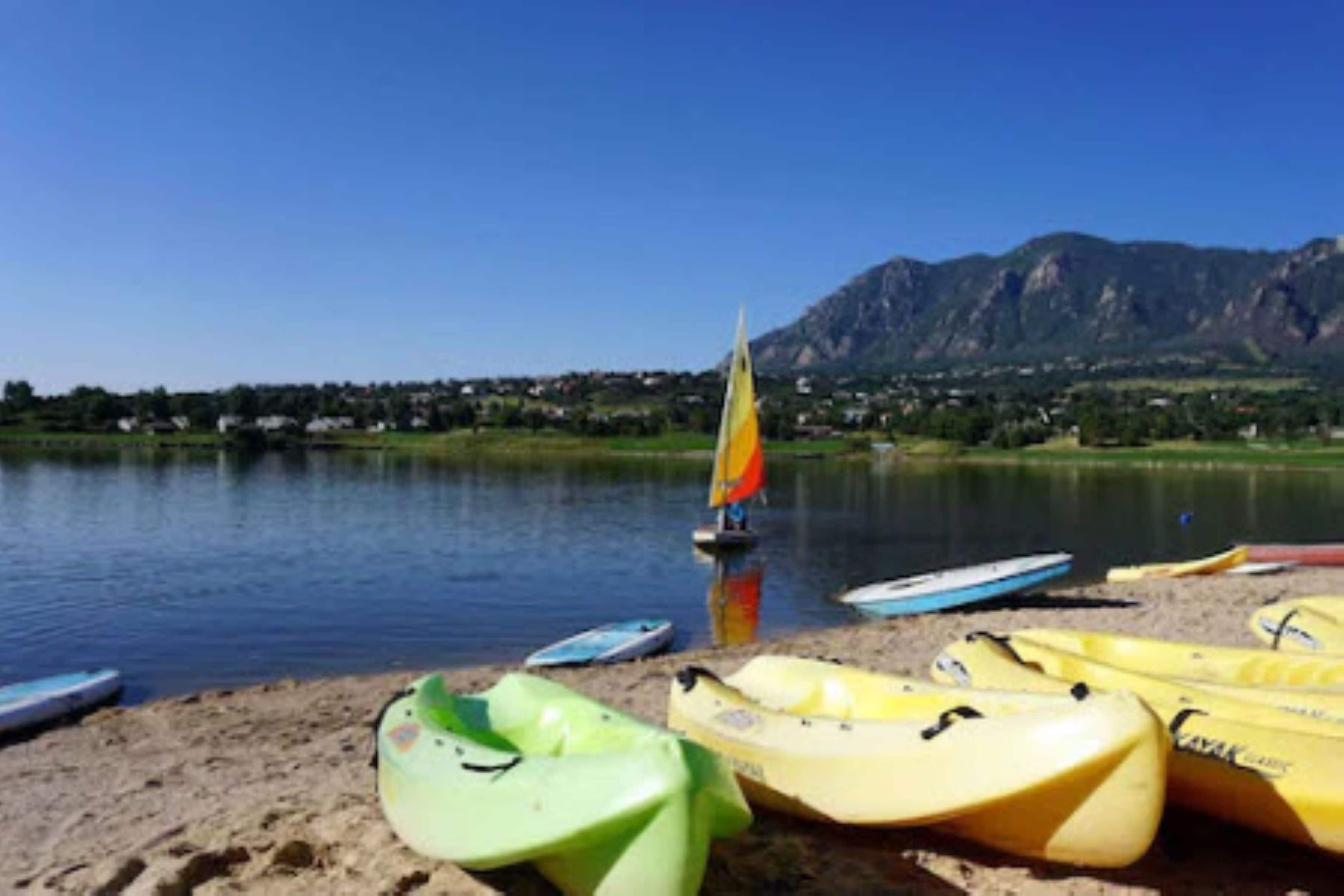A lakeside view with kayaks and a sailboat on the shore, surrounded by mountains.