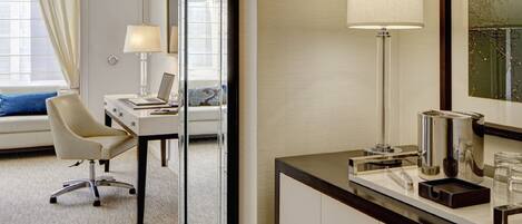 A hotel room with a marble floor, a desk with a laptop, a chair, a large mirror, a bedside table with a lamp, and a bed with a blue pillow.