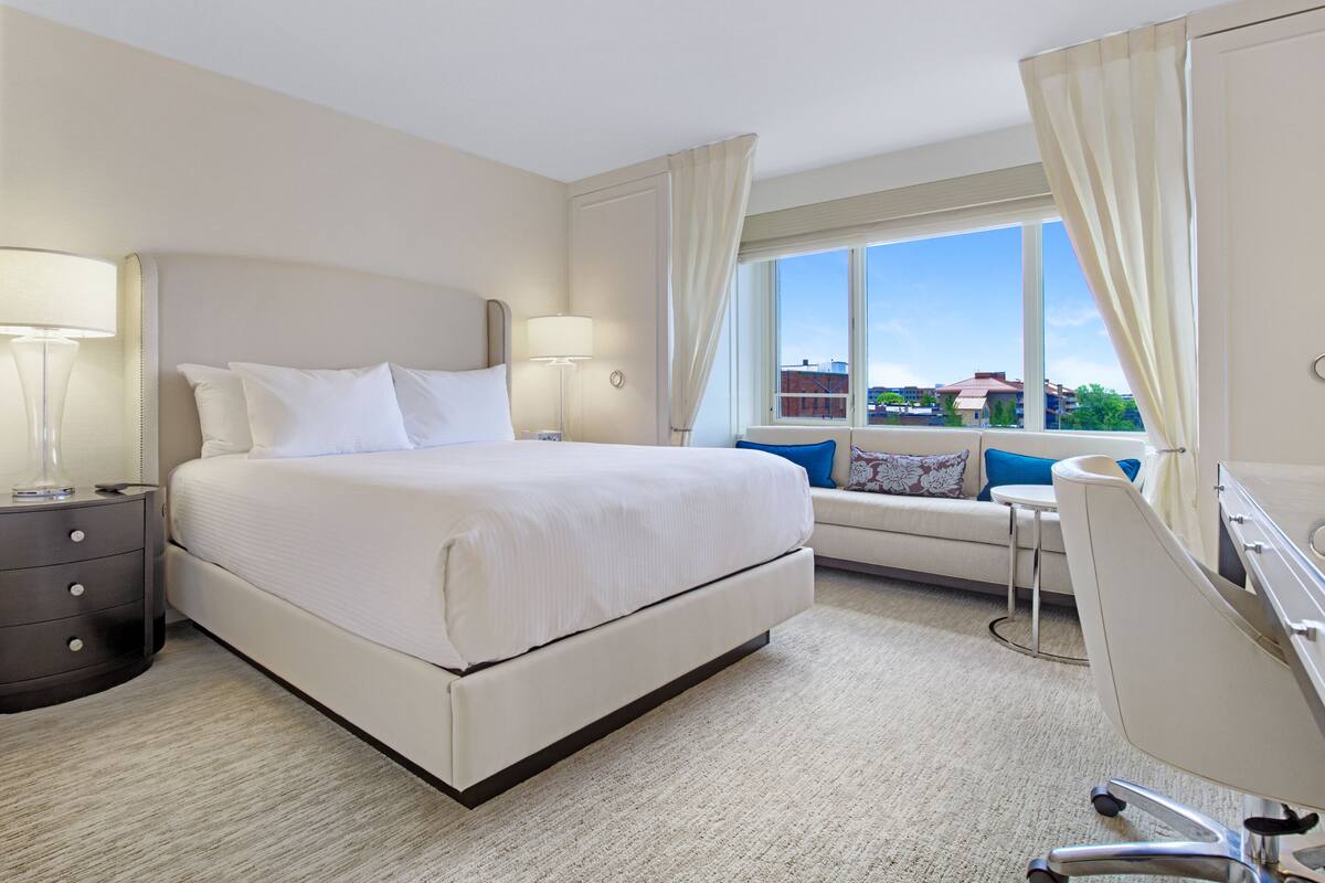 A hotel room with a large bed, bedside lamps, a desk, and a view of buildings through the window.