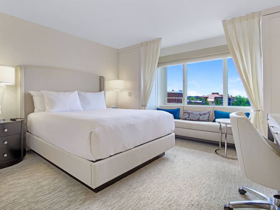 A hotel room with a large bed, bedside lamps, a desk, and a view of buildings through the window.