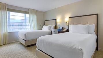 A hotel room with two beds, a window with curtains, a bedside table with a lamp, and a wall-mounted phone.