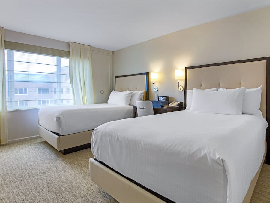 A hotel room with two beds, a window with curtains, a bedside table with a lamp, and a wall-mounted phone.