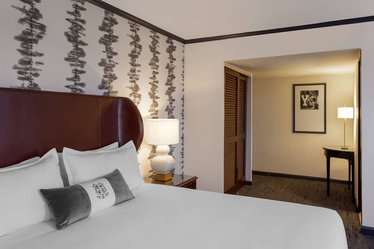 Una habitación de hotel con una cama grande, una mesita de noche con lámpara, un cuadro en la pared y una puerta que da a otra habitación.