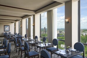 Un comedor con mesas y sillas, ventanas amplias con cortinas, y vistas a un paisaje urbano.