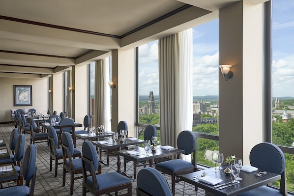 A dining area with tables and chairs, large windows with curtains, and a view of a cityscape.