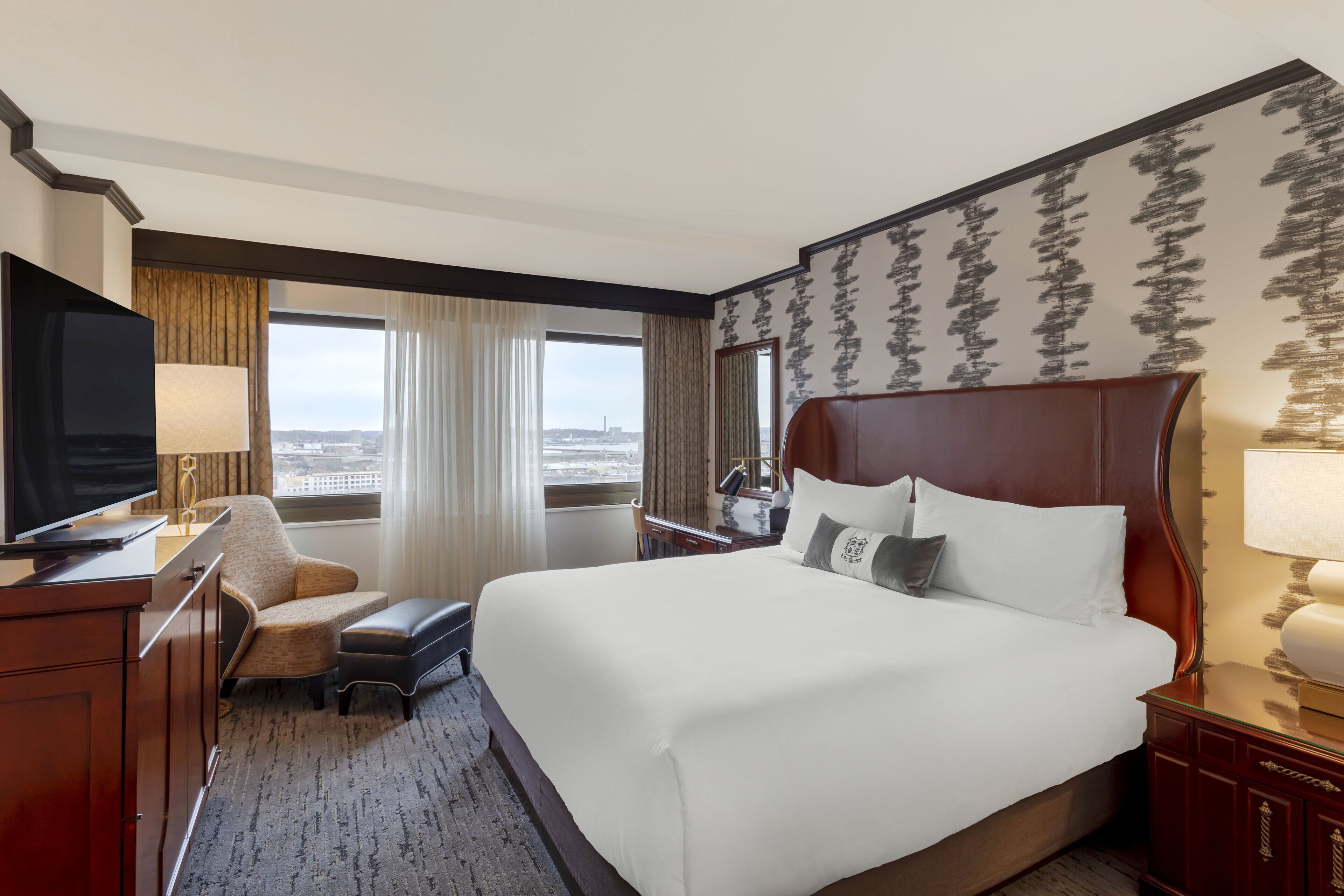 A hotel room with a large bed, a television, a desk, and a view of the city.