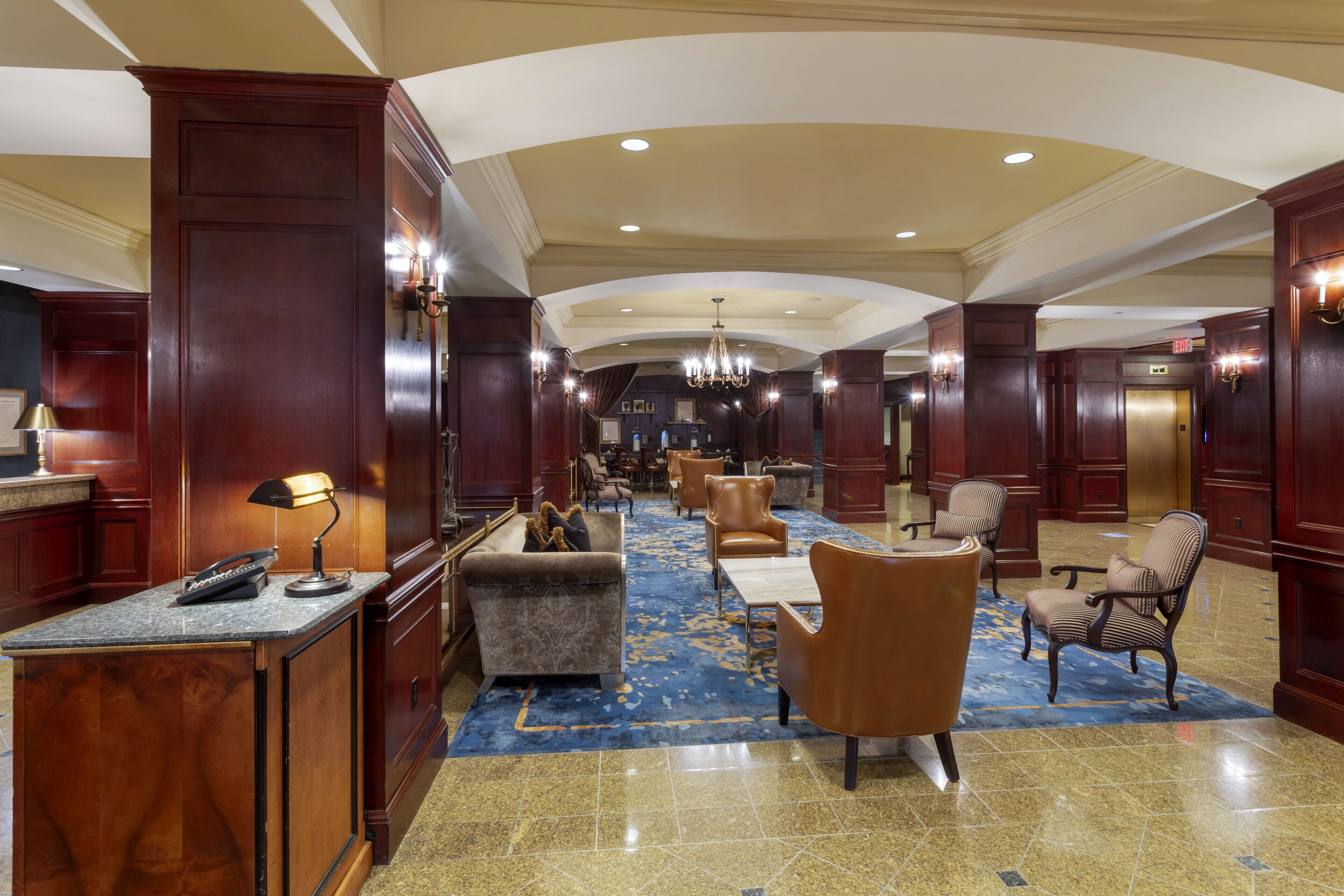 Un hall d’hôtel avec un long comptoir d’accueil, plusieurs fauteuils, une moquette à motifs et un lustre.