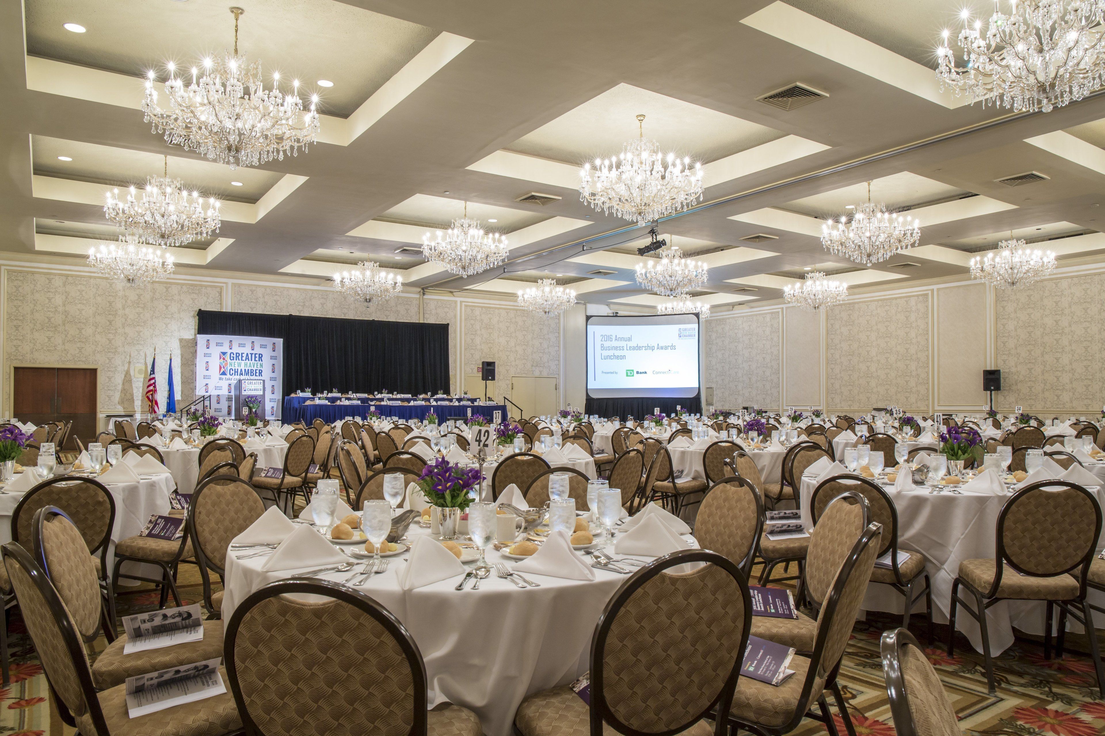 Un salón de banquetes con varias mesas redondas dispuestas para un evento formal, con candelabros y una pantalla de proyección al fondo.