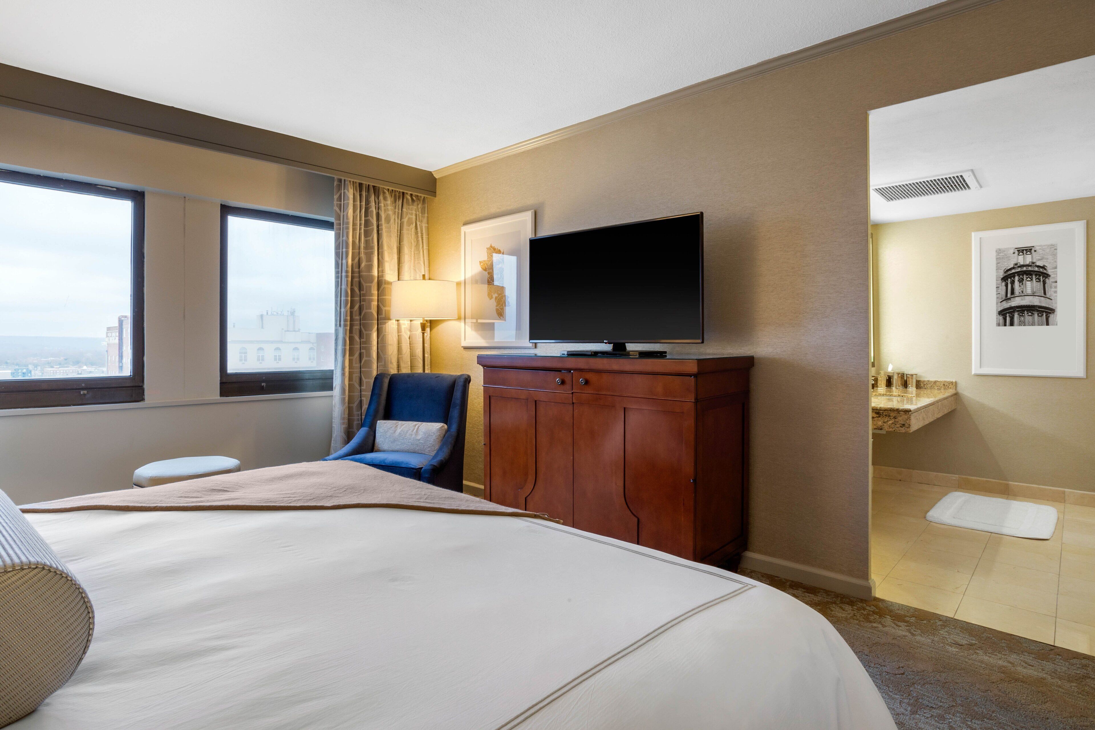 Ein Hotelzimmer mit einem großen Bett, einem Fernseher, einem Sessel und Blick auf die Stadt.