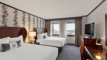 A hotel room with two beds, a TV, a desk, and a view of the city.