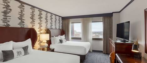 A hotel room with two beds, a TV, a desk, and a view of the city.
