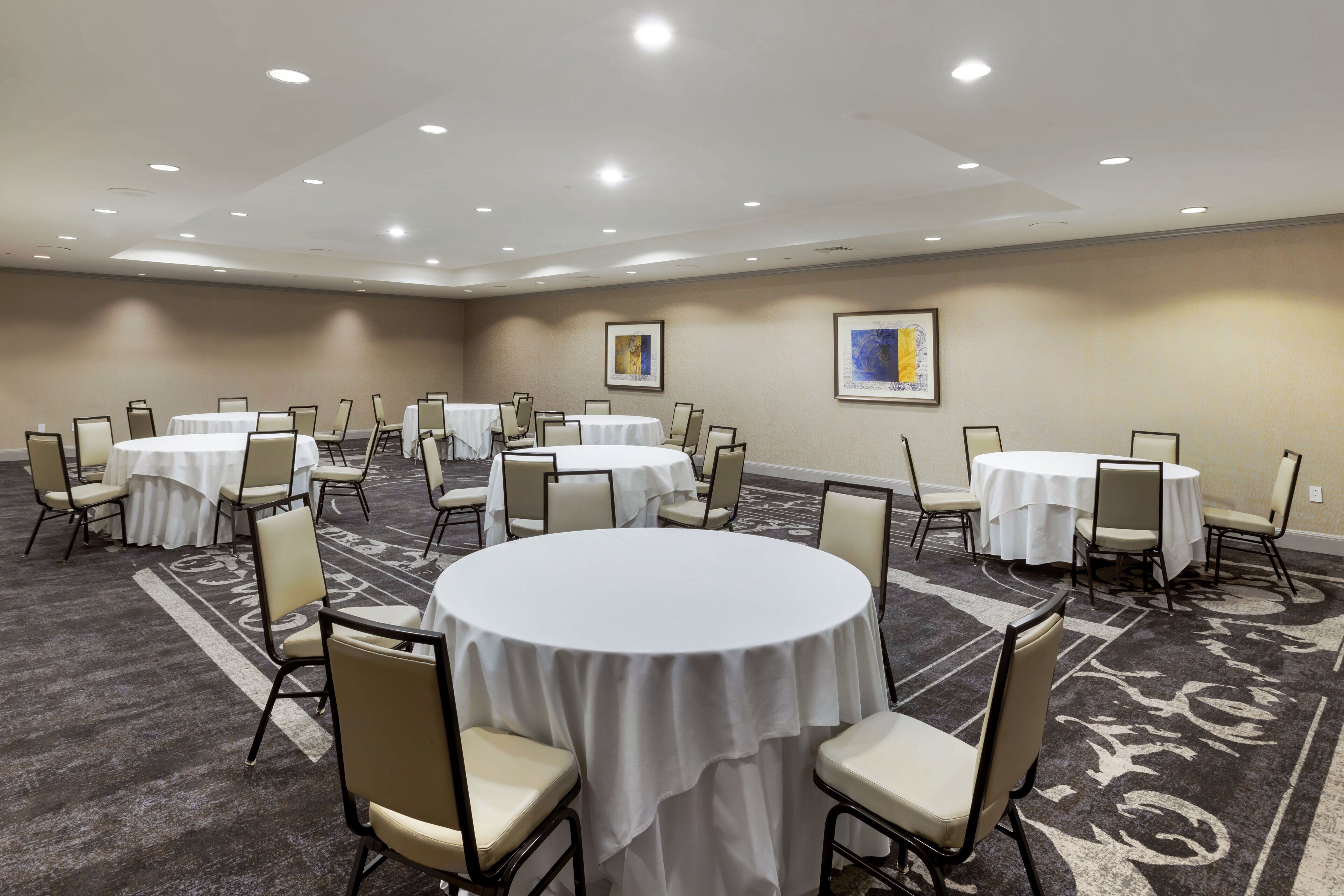 A conference room with multiple round tables and chairs, carpeted floor, and recessed ceiling lights.