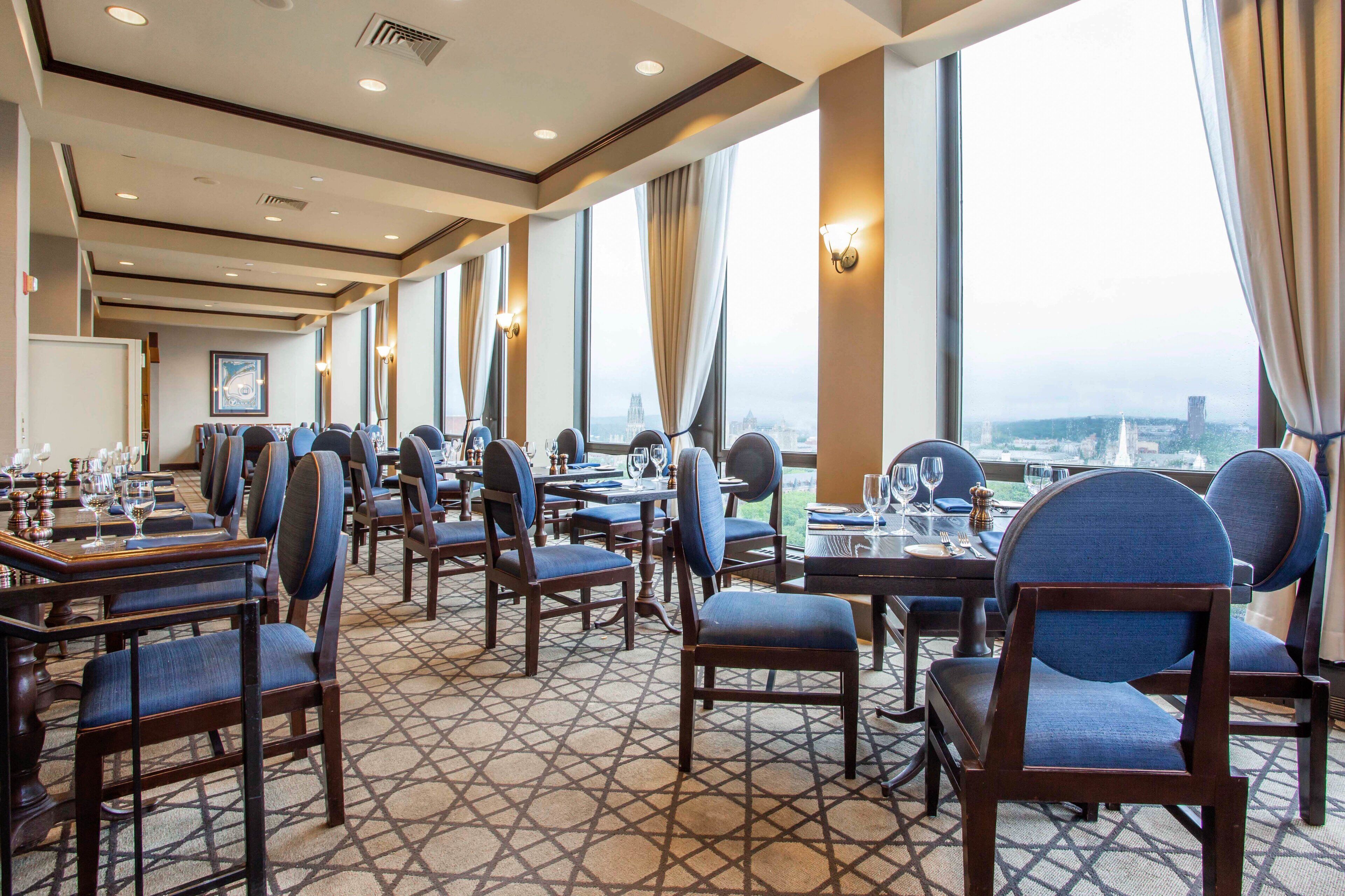 A hotel dining area with multiple tables and chairs, large windows with curtains, and a patterned floor.