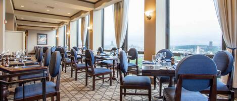 A hotel dining area with multiple tables and chairs, large windows with curtains, and a patterned floor.