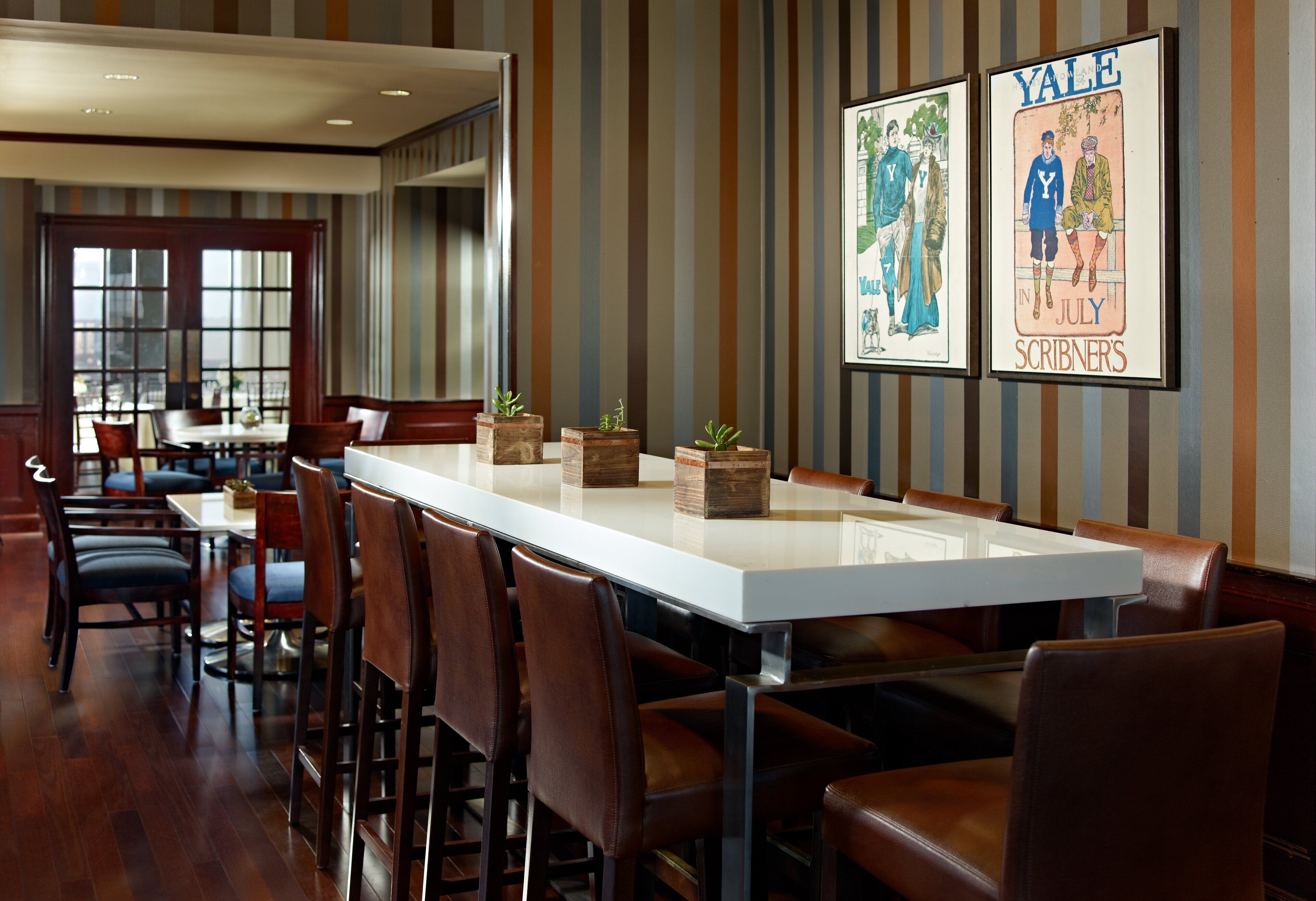 A dining area with a long table and chairs, framed posters on the wall, and a view through a large window.