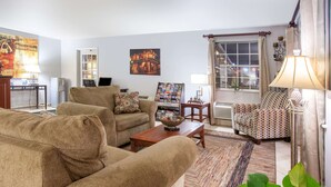 A living room with a sofa, armchair, and coffee table. There is a window with curtains, a lamp, and a poster on the wall. - Super 8 by Wyndham Monroe West (West Monroe)