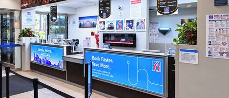 A hotel reception area with a digital display board, a guest services sign, and a cityscape mural on the wall.