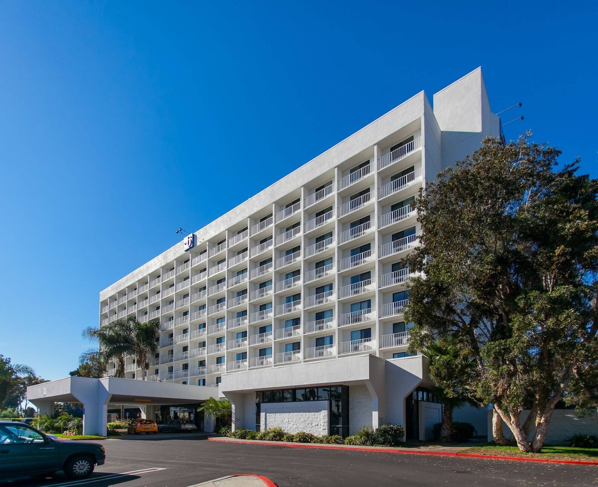 A multi-story hotel with a modern design, featuring balconies on each floor, a central entrance, and a parking area in front.