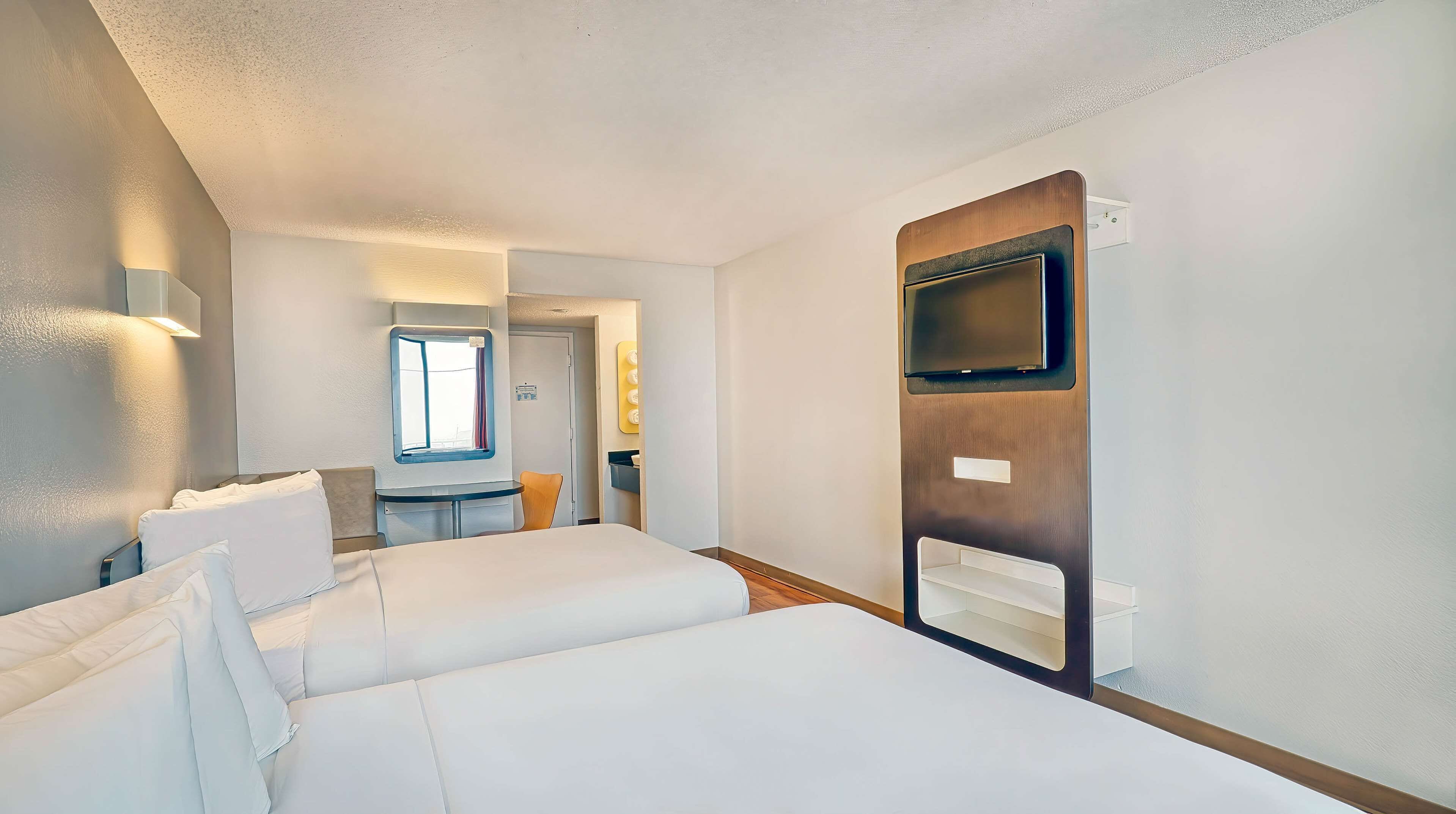 Quarto de hotel com uma cama grande, televisão fixada na parede e uma mesinha com uma cadeira.