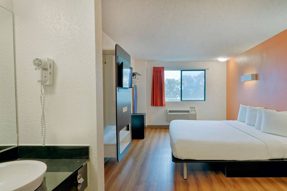 A modern hotel room with a bed, bedside table, and a bathroom with a sink and mirror.