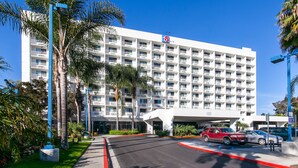 A multi-story hotel with a white facade, balconies, and a parking area with several cars. - Motel 6 Los Angeles, CA - Los Angeles - LAX (Inglewood)