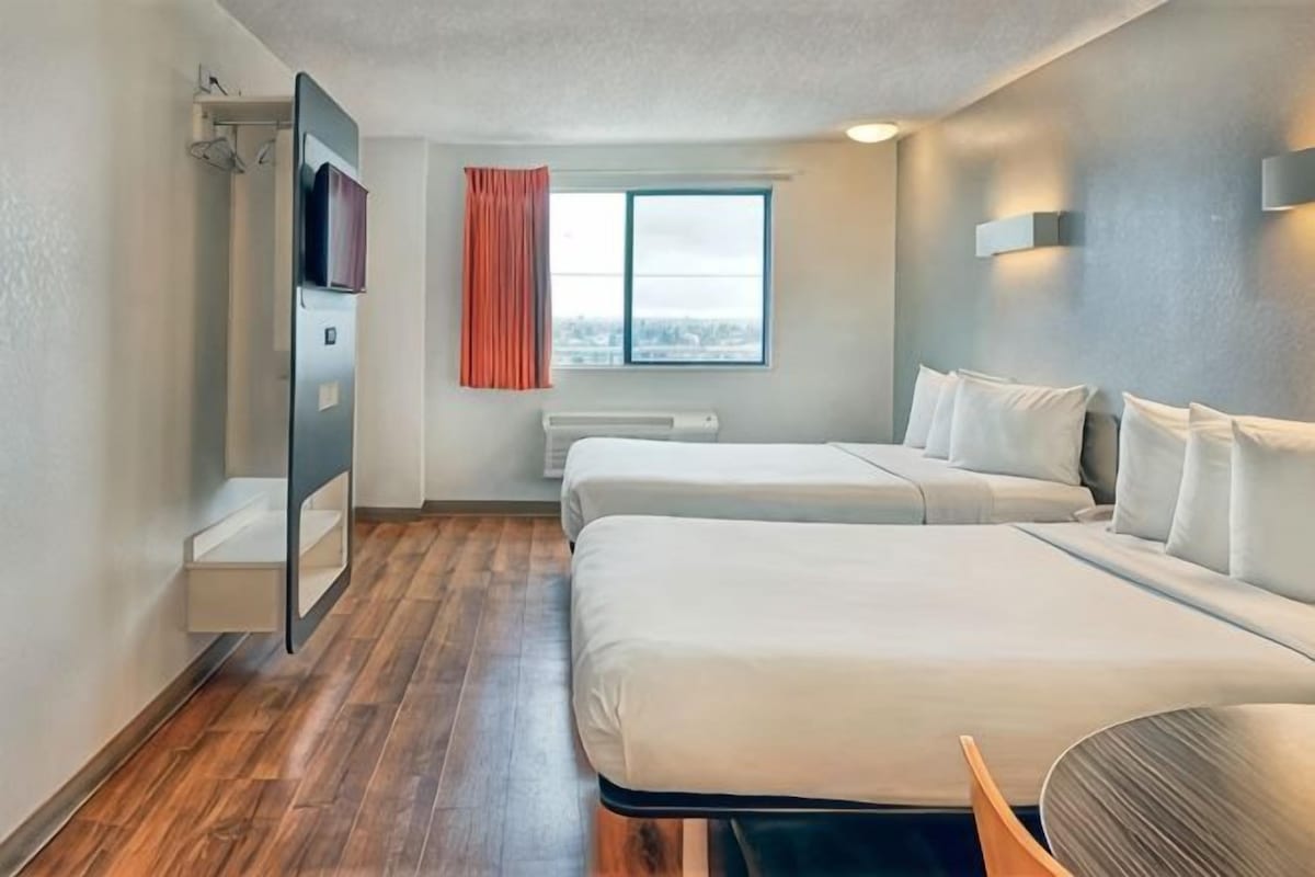 A hotel room with two beds, a window with curtains, a built-in shelf, and a view of the city.