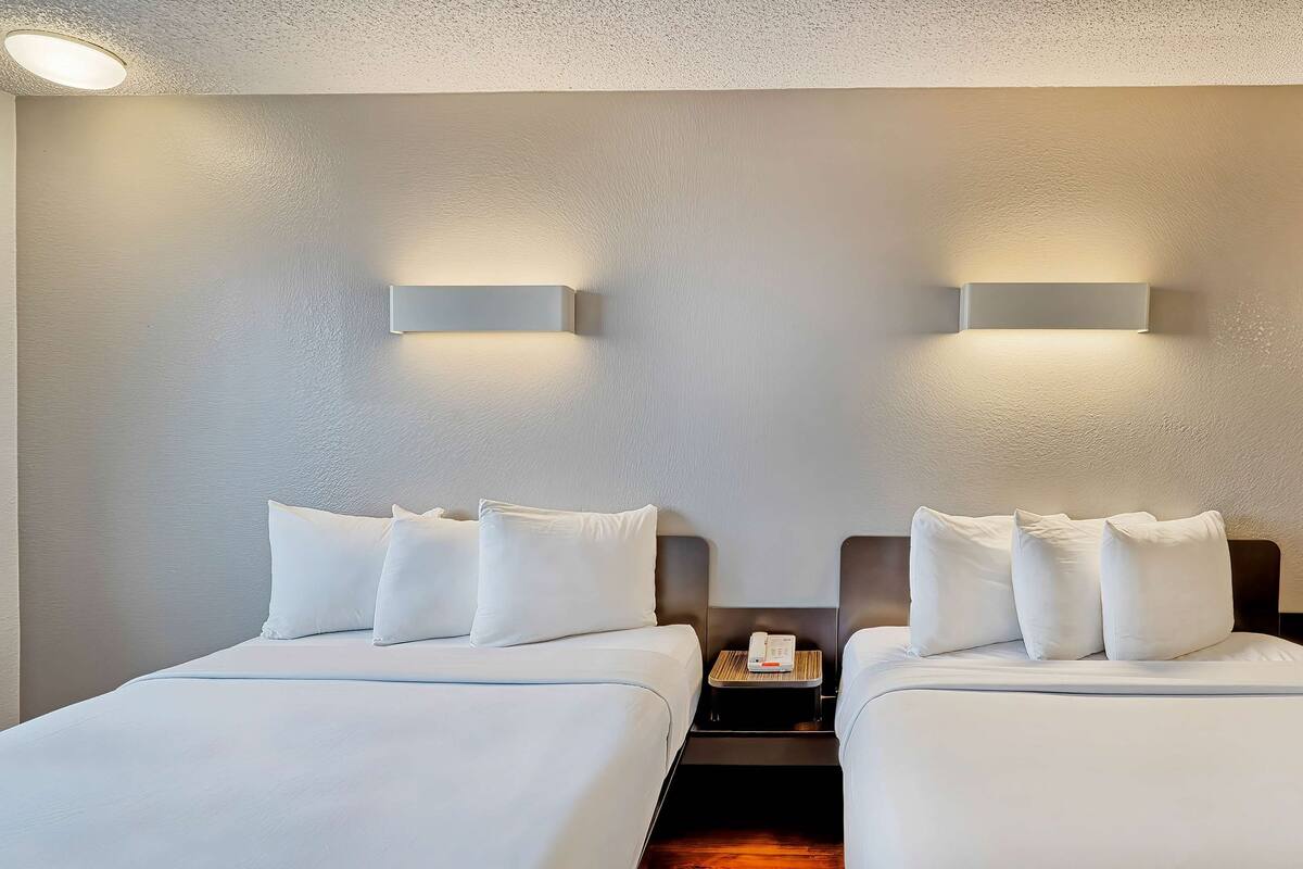 A hotel room with two beds, each with white linens and pillows, a bedside table with a phone, and wall-mounted lighting.