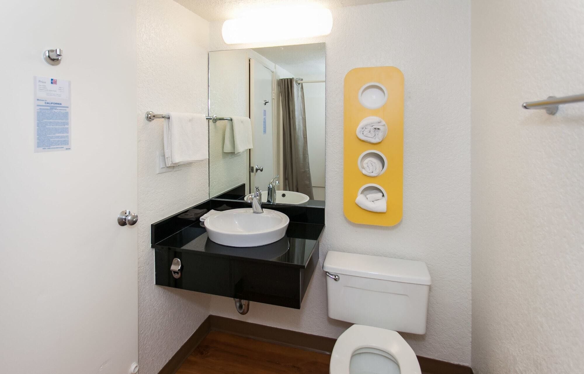 Un baño con un lavabo negro, inodoro blanco y un espejo sobre el lavabo. Se observa un perchero con una toalla blanca colgada.