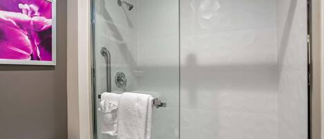 Ducha con cerramiento de vidrio, una toalla blanca colgada en un perchero, un inodoro y un cuadro en la pared.